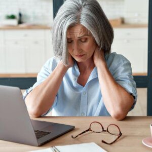 Photo of woman wearing a blue shirt, working at a desk with a laptop.  She's holding her neck because she has neck pain. 